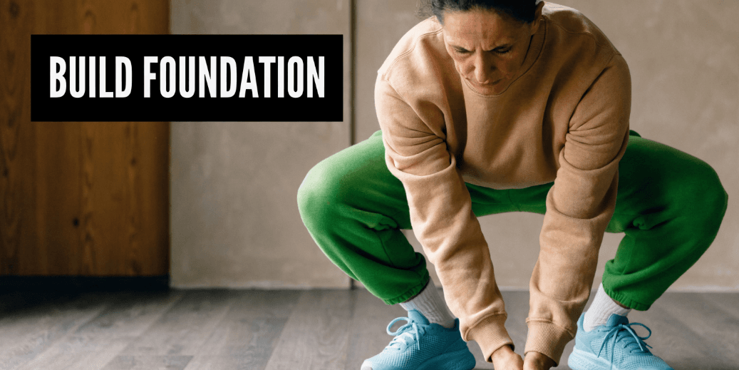 A woman in her forties wearing green sweatpants and a beige sweatshirt performing a deep squat exercise